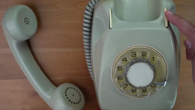 Rotary classic telephone on a wood table. A hand is touching the hang button several times. Off-hook handset.