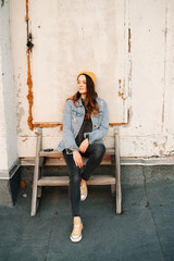 white European woman is sitting on a wooden staircase on the roof of a multi-storey building in stylish denim clothing and a Trendy yellow hat .in the rays of the setting sun. lifestyle concept.