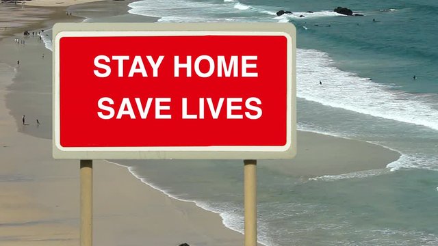 Coronavirus Covid-19 virus pandemic advice to stop the spread of infection. Stay Home Save Lives sign above a beach seashore with a few bathers and surfers. Self isolate.  Corona covid.