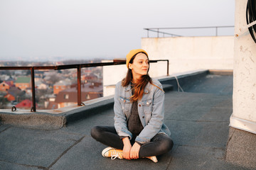 Fototapeta premium pretty white European woman is sitting on the roof of a multi-storey building in a blue denim jacket in gray pants,a yellow stylish hat.in the evening at sunset. lifestyle concept