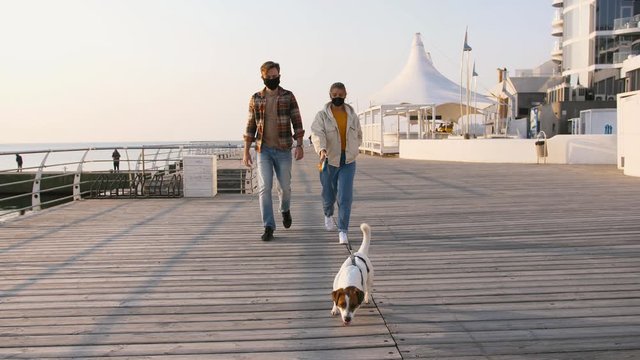 Young happy couple in protective medical masks walking with cute Jack Russel terrier dog outdoors near the sea, slow motion
