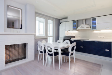 Beautiful white dining area and kitchen with massive dining table and six chairs for with window and modern electric fireplace with mirror interior. Light home relaxation interior concept.