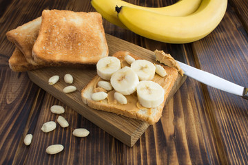 Toasts with peanut paste and banana on the cutting board on the brown wooden background. Closeup.
