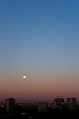 Fototapeta premium Full moon on clear sunrise sky and buildings under it. Blue and pink colours.
