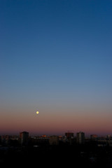 Full moon on clear sunrise sky and buildings under it. Blue and pink colours.