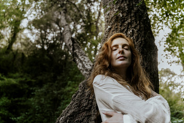Young redhaired woman with closed eyes leaning on tree trunk