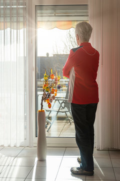 Senior woman standing at closed balcony and looking out of the window