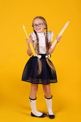 Creative cool first-grader girl with long braids in a school uniform and glasses. In the hands of large pencils. Yellow background. Stationery.