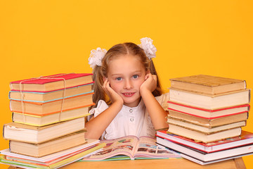 Portrait of a first-grader girl sitting at a desk between stacks of textbooks on a yellow background