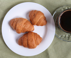 Three fresh croissants on a white plate on a linen tablecloth and a cup of coffee. view from above.