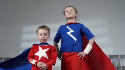 Confident girl and her cute little brother in superhero costumes with capes waving in the wind standing with arms crossed and looking at camera while posing at home