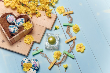 Fototapeta premium Heart shaped box with decorated Easter eggs, yellow flowers, burning candle and brown boards on wooden blue background. Spring and Easter concept. Copy space.