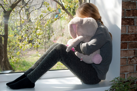 The child is quarantined in isolation due to COVID-19. A child with a soft toy sits on a windowsill and looks out. The concept of quarantined children.