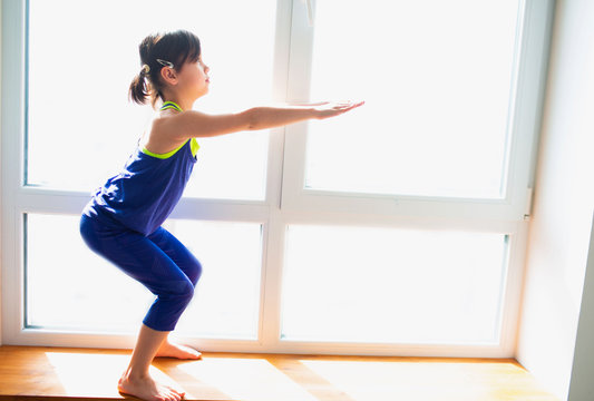 Little girl is doing squat exercises workout at home. Cute kid is training on a wooden windowsill indoor. Little dark-haired female model in sportswear has exercises near the window in her room
