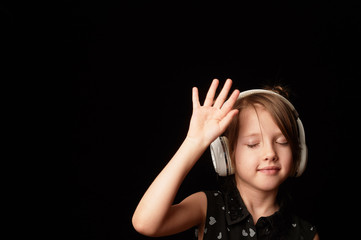 A girl of 6 years, on a dark background. Dancing in white wireless headphones. Hands up. Enjoying the dance.