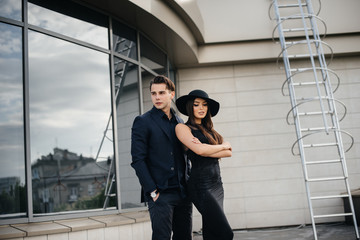 A beautiful, stylish pair of young people in black clothes and glasses stand against the background of an office building in the sunset. Fashion and style