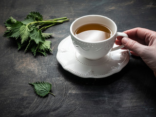 Woman’s hand holding Fresh white ceramic cup with fresh nettle leaves on wooden vintage table.