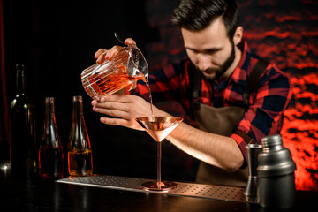male bartender neatly pours cocktail from mixing cup into metal martini glass.