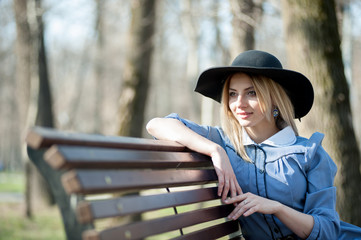 a beautiful blonde girl in a blue dress and a black hat is sitting on a bench and enjoying the spring