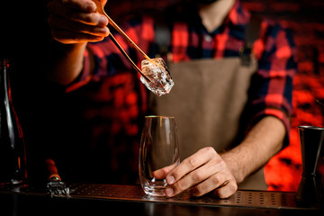 Male bartender put to glass large piece of ice