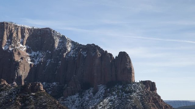 A long-lens timelapse looking up at the snow-covered Shuntavi Butte and Timber Top Mountain in the Kolob Canyons section of Zion National Park. Clouds flow over blue skies in the distance.