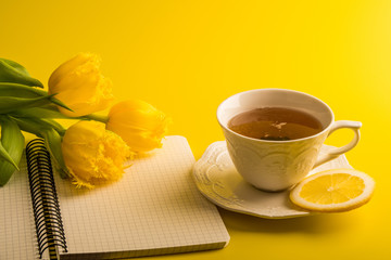 Fototapeta na wymiar Yellow tulips with cup of tea, notepad on yellow