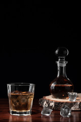 Crystal decanter or carafe of whiskey on cross section of birch tree trunk and glass with alcoholic drink and ice on wooden table in restaurant. Black background for copy space