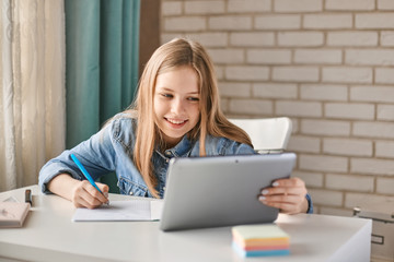 Cute teen schoolgirl does her homework with a tablet at home. The child uses gadgets for learning. Education, distance learning, home schooling during quarantine