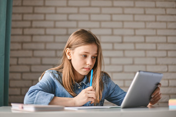 Cute teen schoolgirl does her homework with a tablet at home. The child uses gadgets for learning. Education, distance learning, home schooling during quarantine