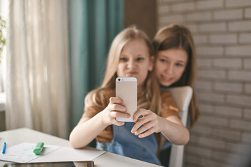 Two sisters take a break from homework and take selfies on their mobile phone while sitting at a table. Education, distance learning, home schooling during quarantine