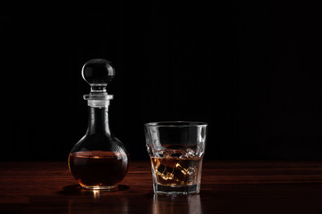 Crystal decanter or carafe of whiskey and glass with alcoholic drink and ice on wooden table in restaurant. Black background for copy space