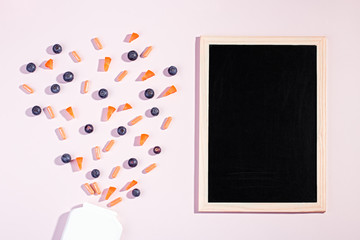 Vitamins and supplements for healthy eyes on pink background. Blueberry, carrot pieces and orange capsules flying out from white bottle with black board for text, flat lay, top view