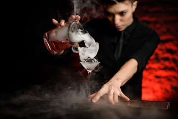 young female barman pours smoky cocktail from mixing bowl into martini glass.