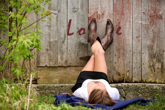 Woman laying on the ground with her cowboy boots on the side of a barn spelling out the word Love.