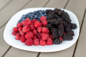 White ceramic plate with assorted sweet juicy organic seasonal berries for snack. Ripe tasty fresh raspberry, blackberry and blueberry summer mix for breakfast morning. Healthy vitamin nutrition diet