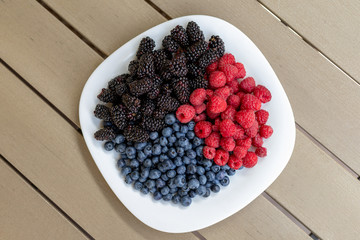 White ceramic plate with assorted sweet juicy organic seasonal berries for snack. Ripe tasty fresh raspberry, blackberry and blueberry summer mix for breakfast morning. Healthy vitamin nutrition diet