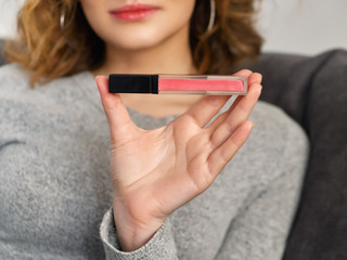 Woman's beauty blogger hands holding professional hi-end cosmetic coral liquid lipstick with blurred body background, warm cozy tones and copyspace