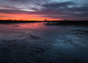 The vivid colors of a winter sunset stream across the frozen surface of a Midwest wetland environment.