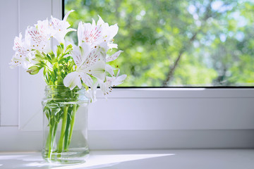 A bouquet of white flowers Alstroemeria stands on a white windowsill under the rays of the sun on a background of trees. The concept of flowers, background, holiday, comfort.