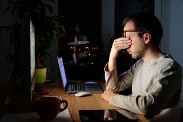 Tired freelancer man rubbing his eyes, sitting at desktop PC/laptop late at night, during the period of self-isolation and remote work at home, falls asleep from fatigue.  