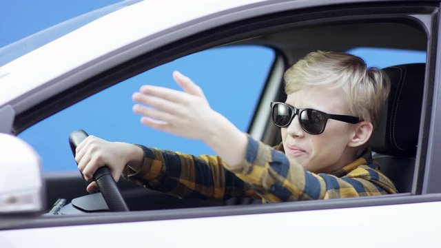 A little shouting blonde boy in casual clothes is travelling. Handsome boy is driving car with greenscreen background. He is sitting in the car. He is aruing with someone