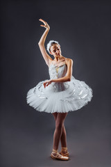 Fototapeta premium A young graceful ballerina dressed in professional attire, pointe shoes and a white tutu, demonstrates dance skills. Beautiful classic ballet dancer isolated on grey backgroung in photostudio