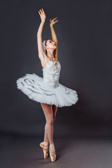 Portrait of young beautiful graceful caucasian ballerina practice ballet positions in tutu skirt of white swan from Swan Lake. Classical Ballet dancer in studio on gray background standing on tiptoe