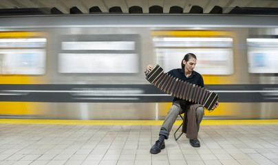 Tango performer playing a bandoneon with hat and subway in motion.