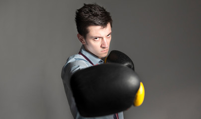 Low key portrait of handsome young businessman wearing boxing gloves and punching towards camera. Selective focus on the face. He is wearing a shirt and tie that is undone. 