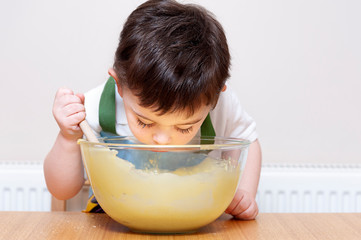 Cute young boy stirring cake mix with a wooden spoon and leaning into the bowl to smell the contents. The mixture is in a glass bowl. He is sat in the kitchen. 