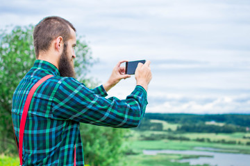 Handsome man hipster or guy with beard and moustache on serious face in cloth shirt and suspenders sunny outdoor on mountain top against cloudy sky on natural background, copy space, makes a photo.