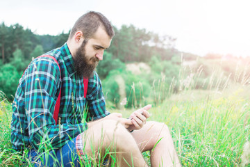 Naklejka na ściany i meble Handsome man hipster with beard on serious face in cloth shirt and suspenders sunny outdoor on mountain top against cloudy sky on natural. Tourism concept. A guy with a phone. Work in nature.