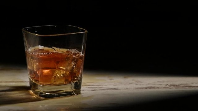 Person pouring ice cubes into glass with bourbon on wooden table in rays against black background