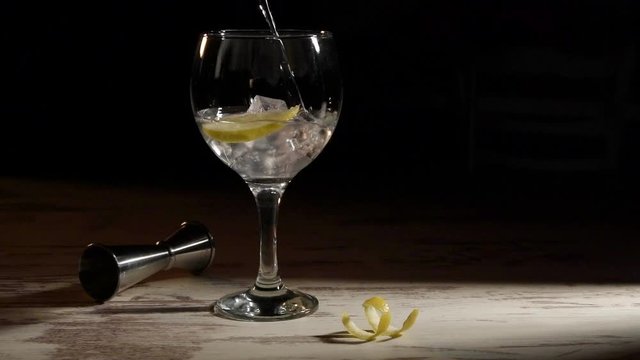 Person filling transparent glass of gin tonic with lemon and jigger on table in dark room
